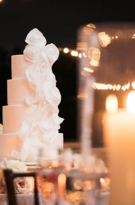 Four tiered white wedding cake with fondant bows on each tier, photographed in the reception tent