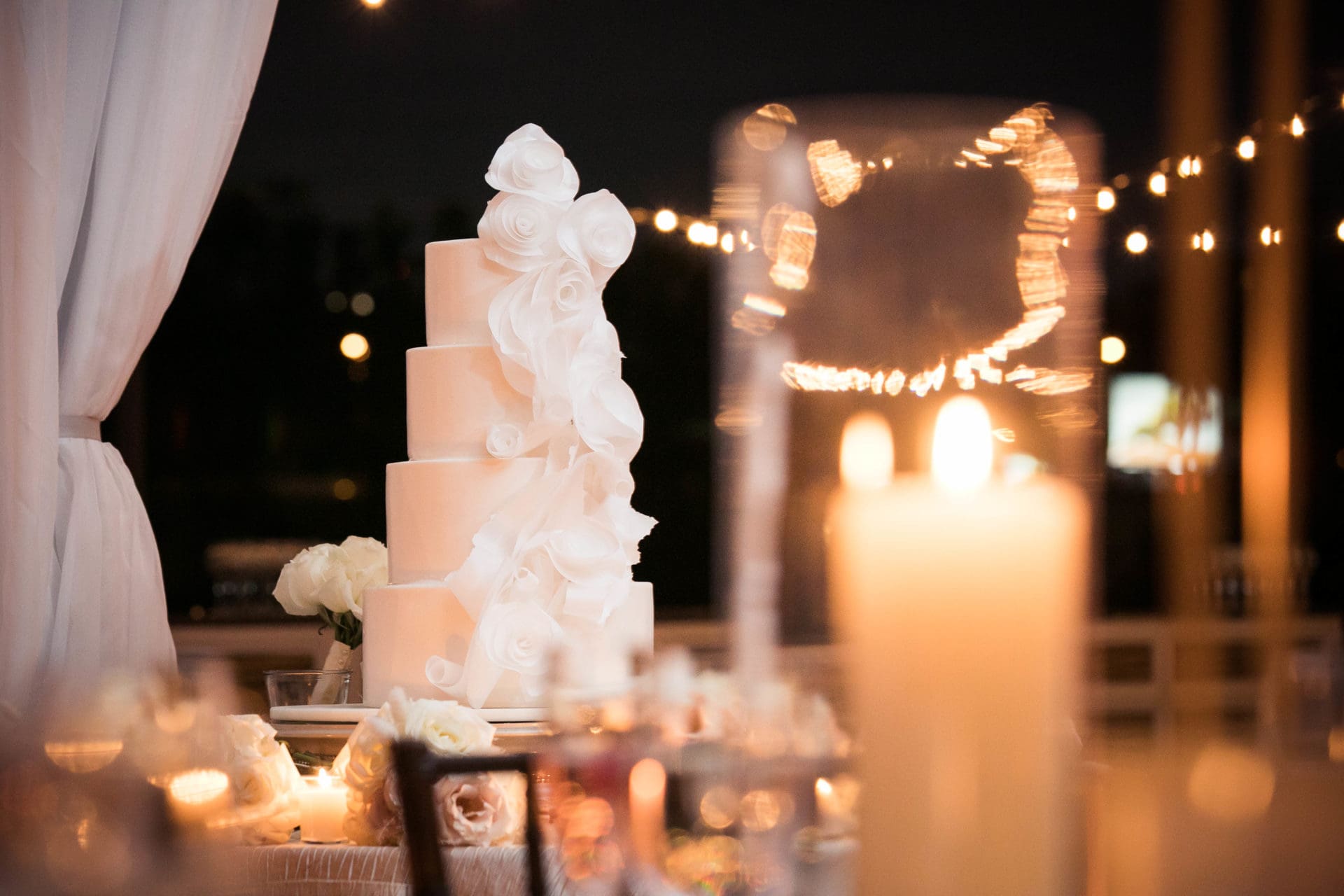 Four tiered white wedding cake with fondant bows on each tier, photographed in the reception tent