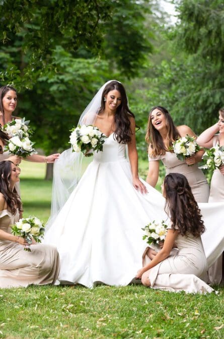 A bride and her bridesmaids looking at each other and laughing on the green lawn in front of Lyndhurst Castle in Tarrytown, NY