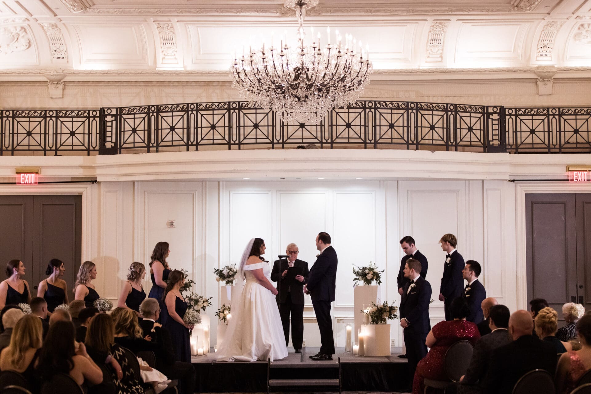 A bride and groom are standing on a stage during their wedding ceremony while the officiant talks to them and their guests