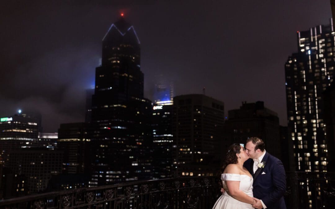 A bride and groom kissing on the balcony at night at the Bellevue Hotel in Philadelphia with the skyline behind them