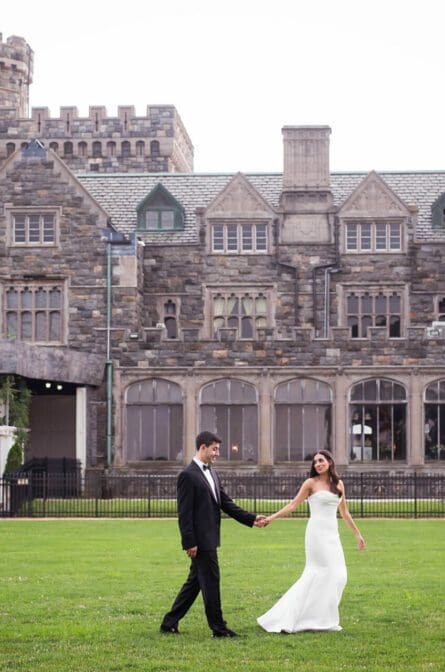 A bride and groom are walking across the front lawn of the Hempstead House for their summer wedding