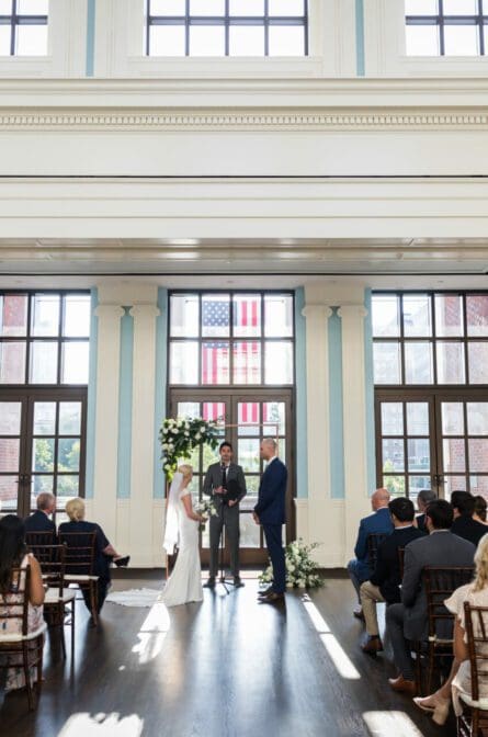 A bride and groom are standing in a large room with floor to ceiling windows and exchanging vows underneath a flower arch