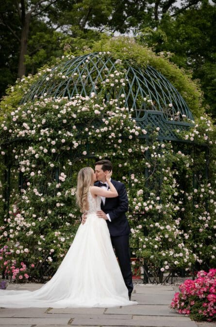 Bride and groom kiss in front of a flower covered gazebo in the garden of the New York Botanical Garden